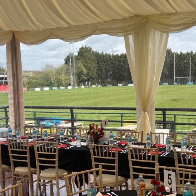 A long table dressed in black linen with red roses, looking out through clear marquee walls to a rugby pitch